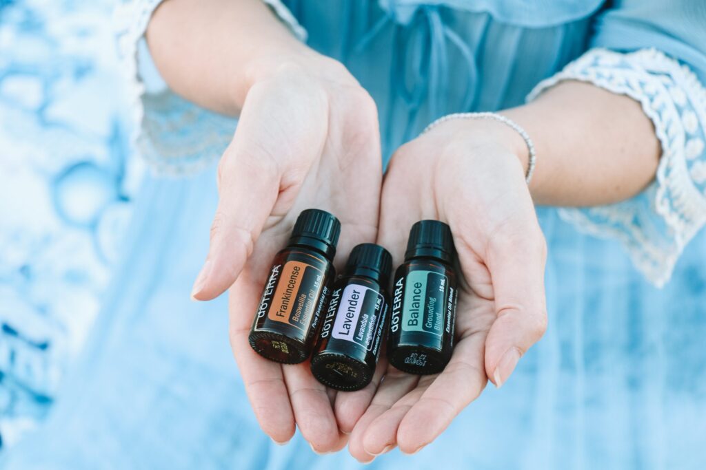 A woman wearing a blue dress holds three bottles of doterra essential oils in her palms, with labels that show their contents: Frankincense, Lavender and Balance, a grounding blend of oils
