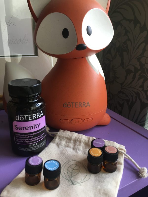 A photograph of a an orange and white fox shaped diffuser on a purple surface, next to a container saying doTERRA Serenity and five essential oil samples onto on a flat unbleached cotton bag.